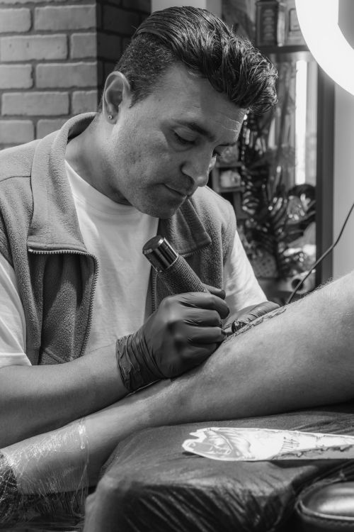 Free Black and white photo of a tattoo artist focused on tattooing a client's leg in a studio. Stock Photo