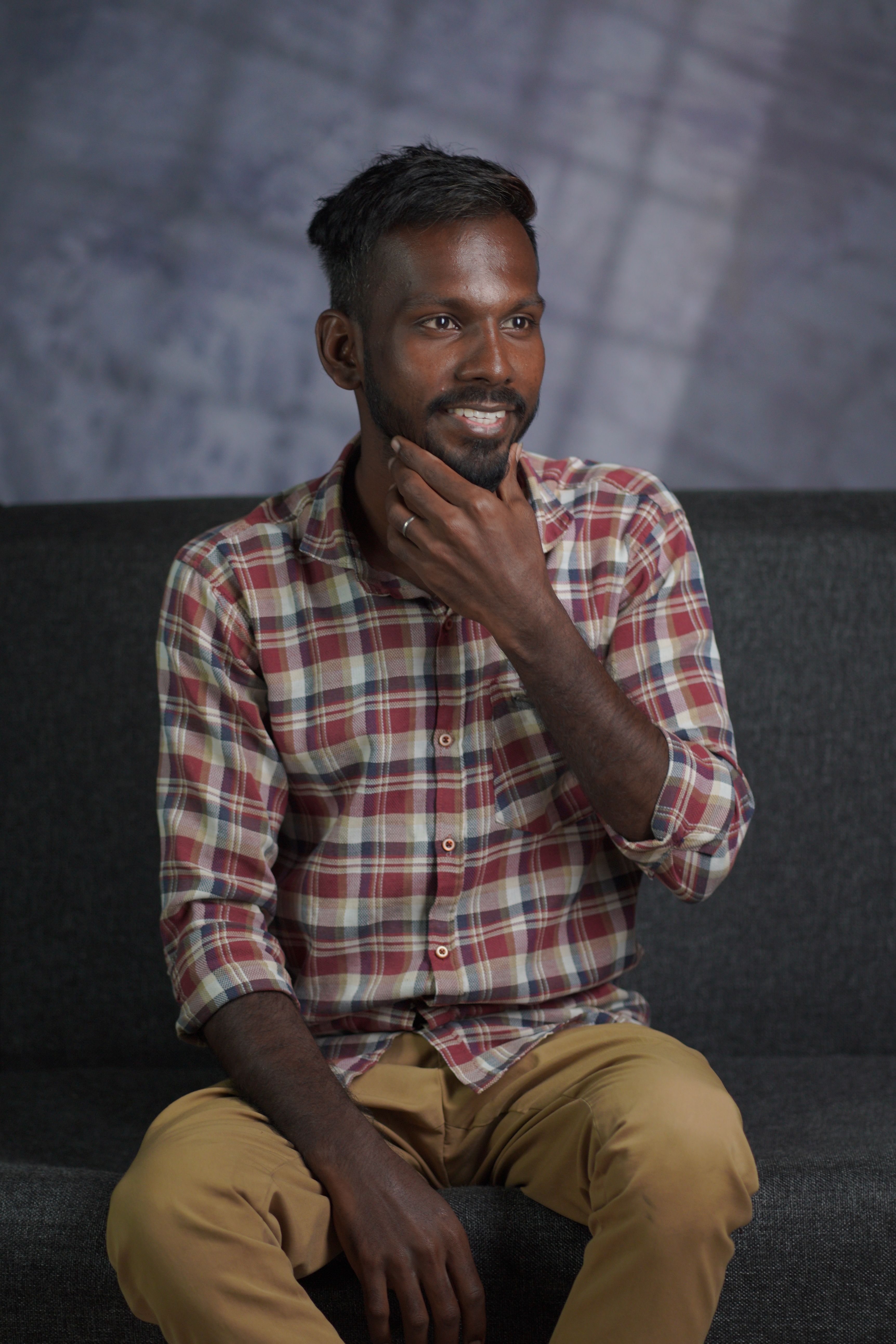 Portrait of a smiling man seated on a sofa, hand on chin, wearing a plaid shirt.