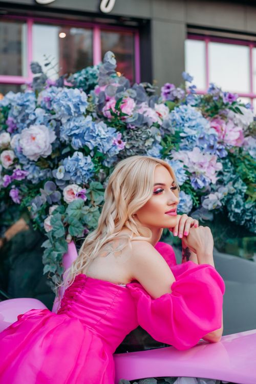Free A fashionable blonde woman in a pink dress poses elegantly in a floral setting outdoors. Stock Photo