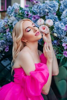 A beautiful woman in a pink dress poses with roses and hydrangeas in a vibrant outdoor setting.