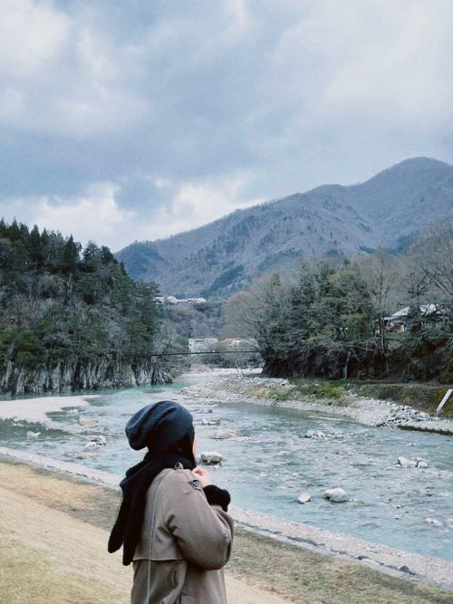 Free A woman in a beanie and coat gazes at a serene river flanked by mountains and trees. Stock Photo