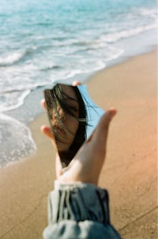 A woman's face reflected in a mirror, held by the sea. Artistic blend of nature and self-discovery.