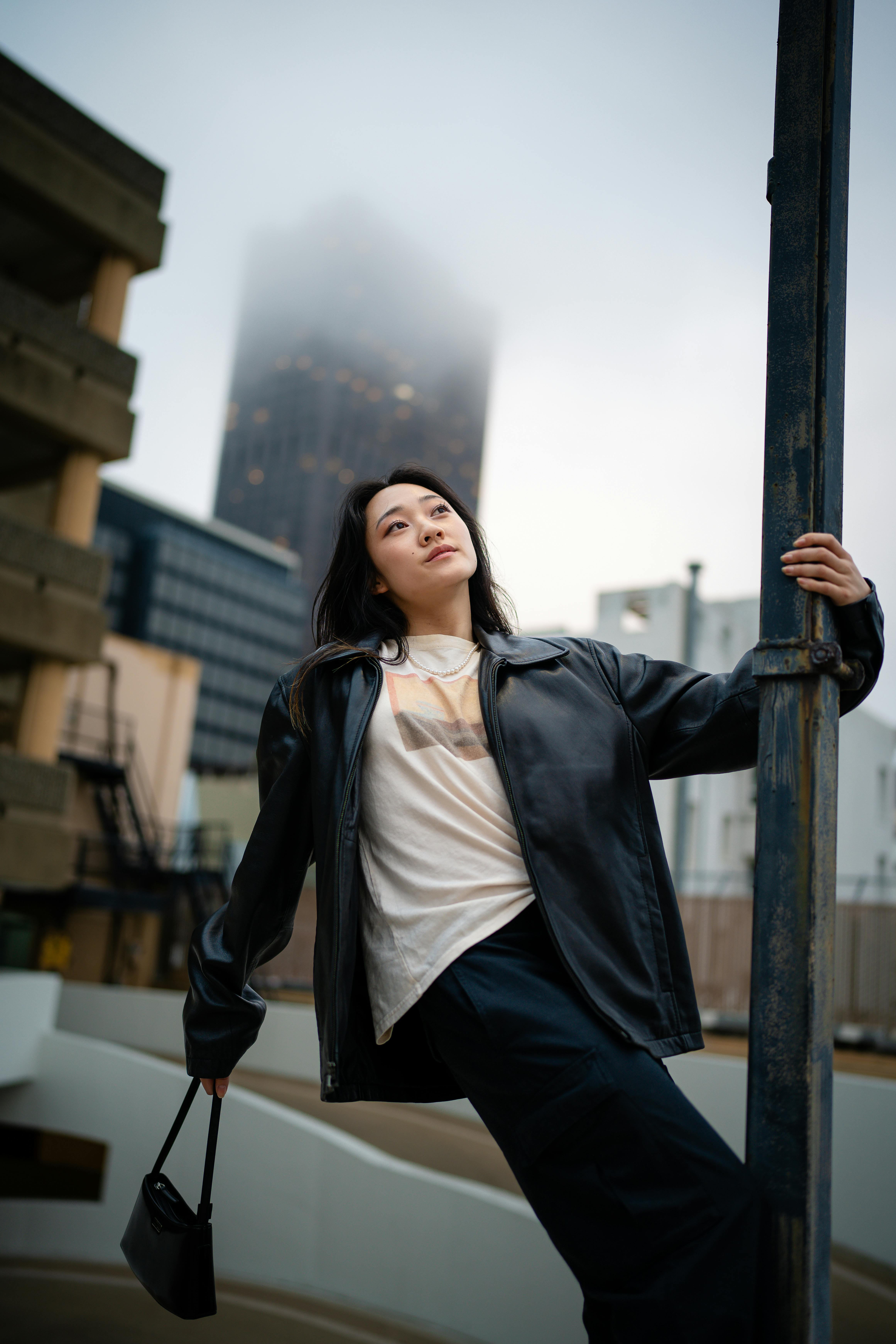 Asian woman in a black jacket leaning against urban post on a foggy day, showcasing modern fashion.
