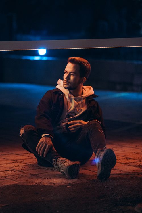 Free Young man sitting on sidewalk, illuminated by city lights, lost in thought during a quiet night. Stock Photo