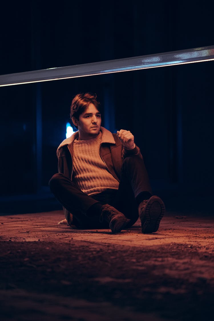 Young Man In A Sweater And Jacket Sitting On The Ground At Night 