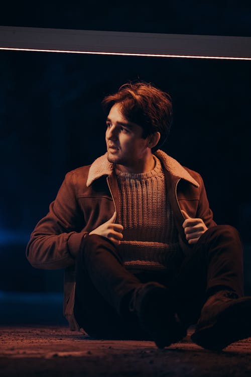 Man in Jacket and Sweater Sitting on Ground in Evening