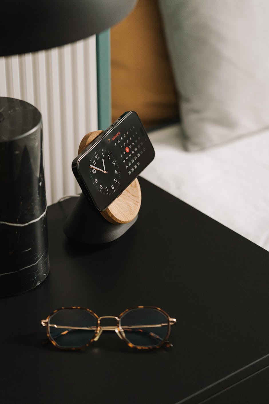 A stylish office desk setup featuring a smartphone docked as a clock with eyeglasses in the foreground.