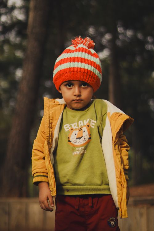 Free A young boy wearing a colorful hat and jacket stands outdoors in a forested area during winter. Stock Photo