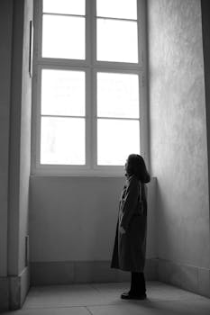 Black and white photo of a woman standing alone, gazing out a large window.