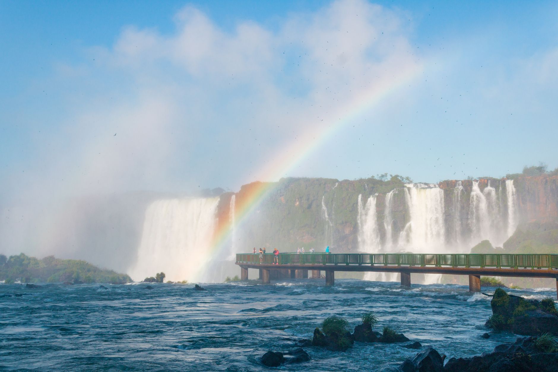 A vivid rainbow arching over a green metal walkway at Iguazú Falls, with curtains of water cascading behind and visitors on the footbridge above the river