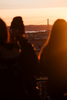 Backlit silhouettes admire a stunning Lisboa sunset with a view of a prominent bridge.