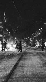 Pedestrians and cyclist navigate a snow-covered street under city lights on a winter night.