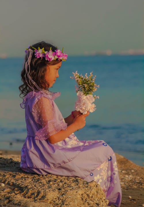 Girl in a Purple Dress Sitting on the Beach with a Bouquet in her Hands 