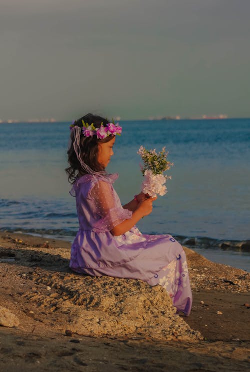 A Girl in a Dress Sitting on the Beach and Holding Flowers