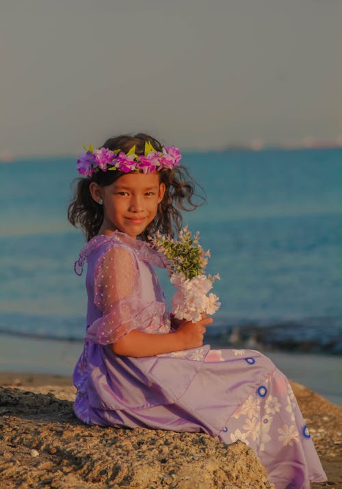A Girl in a Dress Sitting on the Beach and Holding Flowers