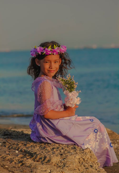 A Girl in a Dress Sitting on the Beach and Holding Flowers