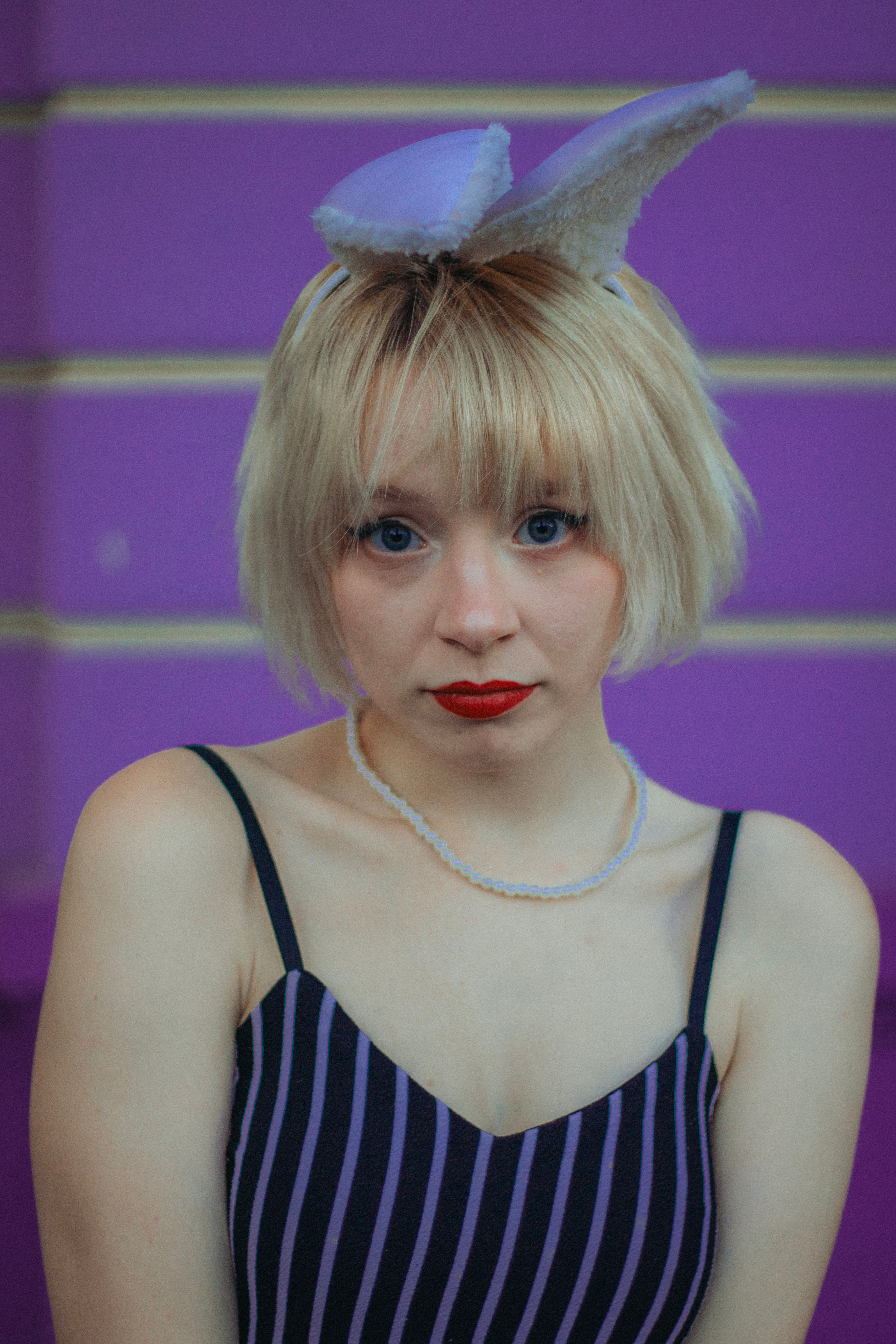A young woman with blonde hair, bunny ears, and red lips posing against a vibrant purple background.