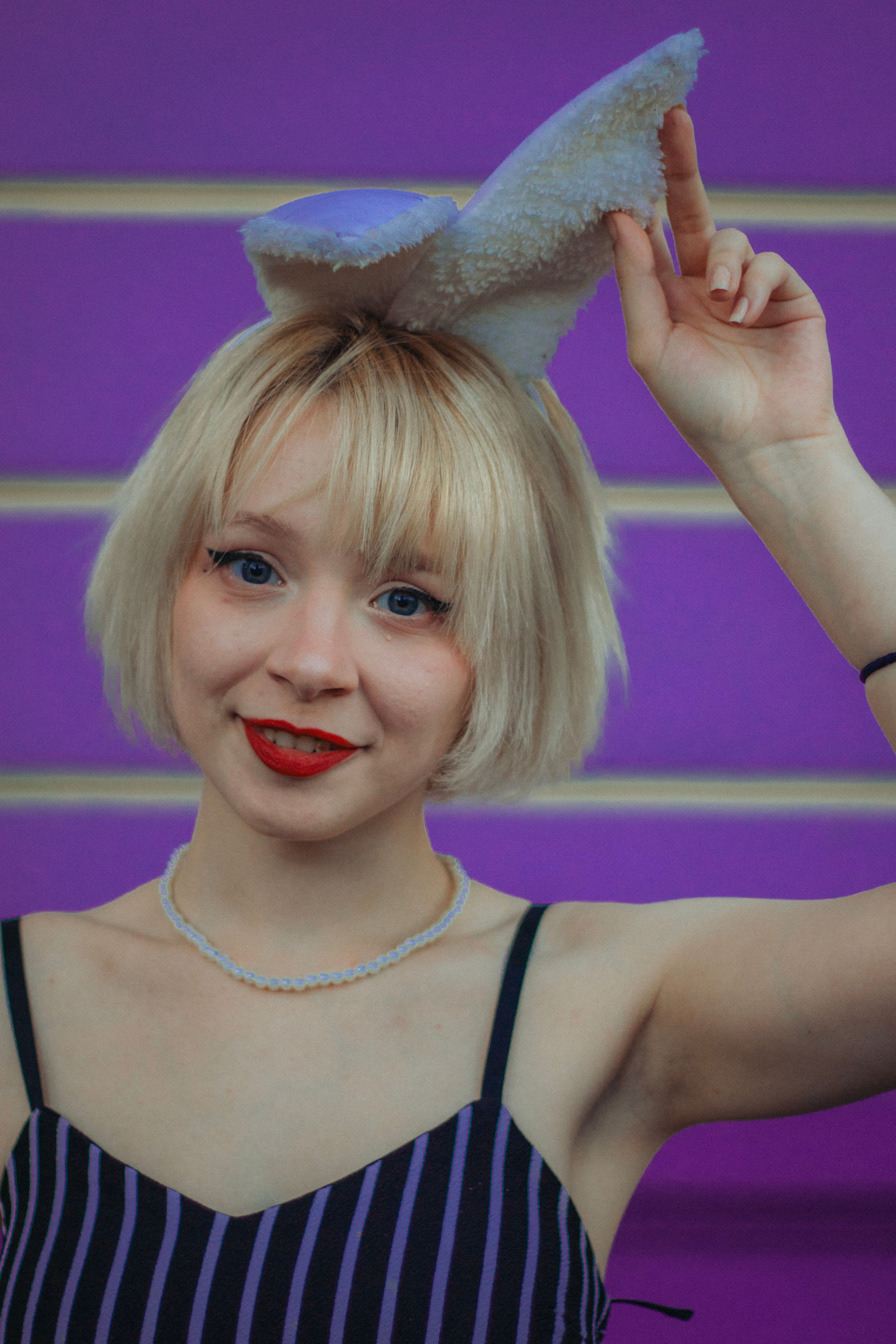 Young woman smiling with bunny ears, red lips, and a necklace against a vibrant purple backdrop.