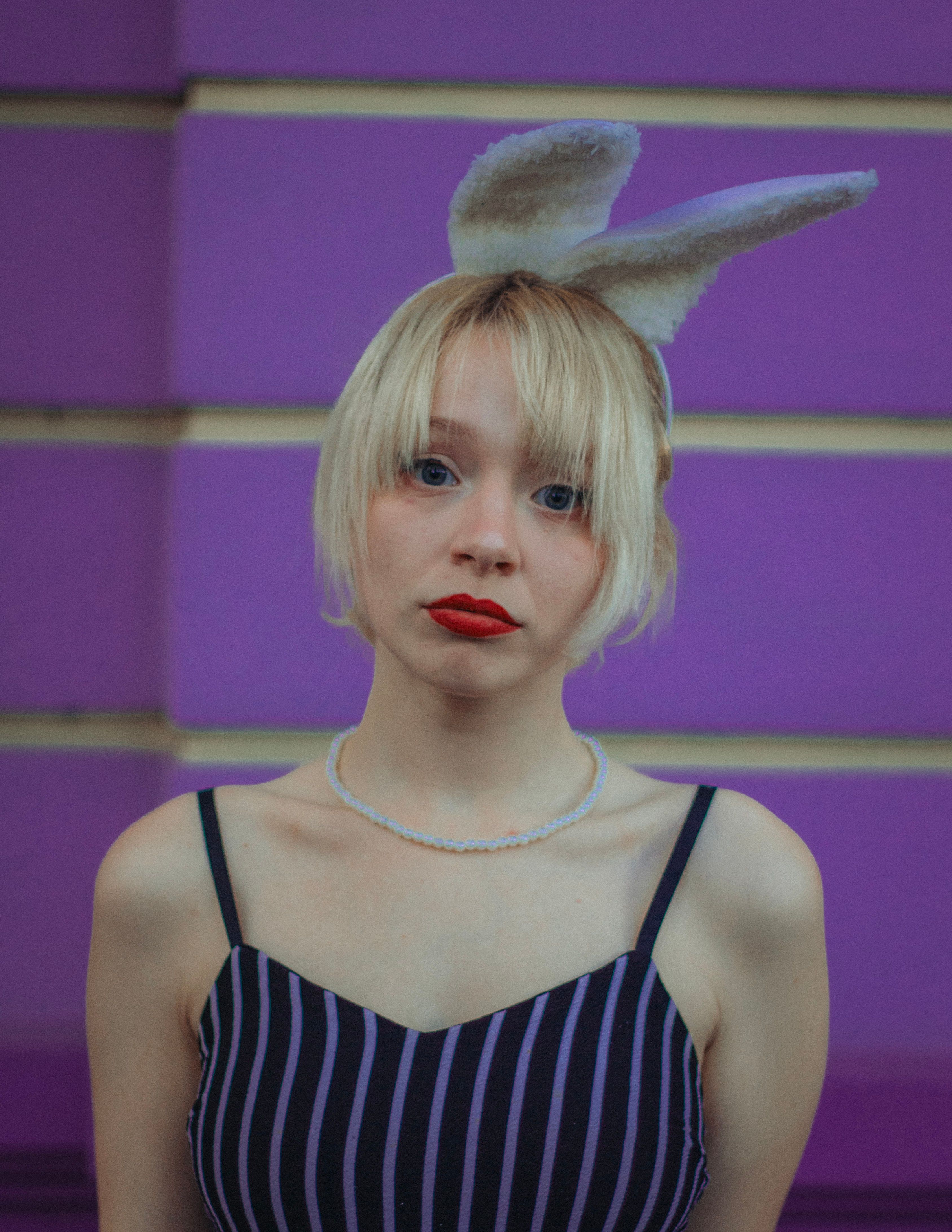 Portrait of a blonde woman wearing a striped dress and bunny ears in front of a purple wall.