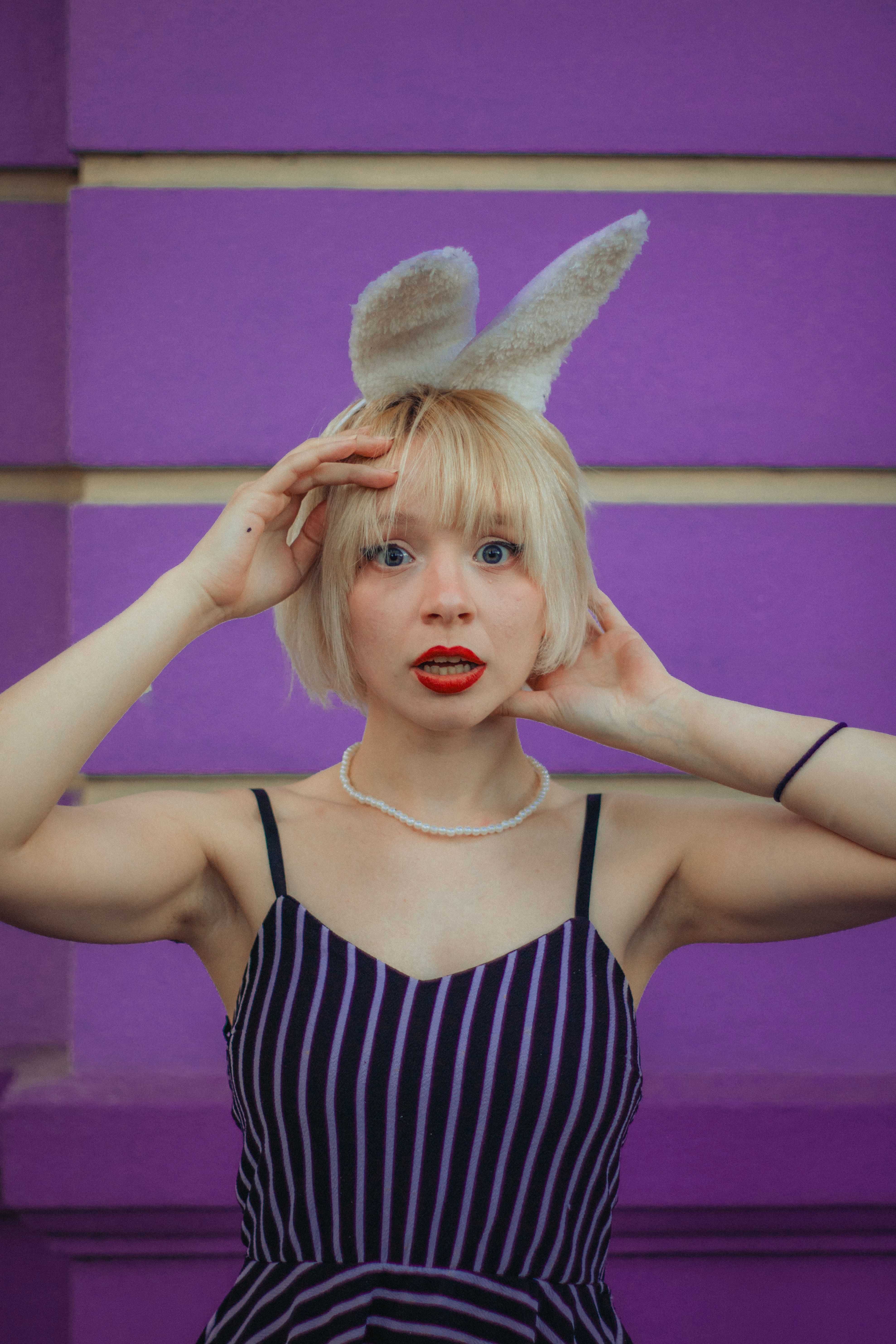 A whimsical portrait of a woman with blonde hair wearing bunny ears and a striped dress against a purple background.