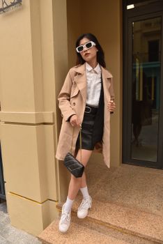 Fashionable woman poses elegantly on a city street wearing a beige coat and sunglasses.