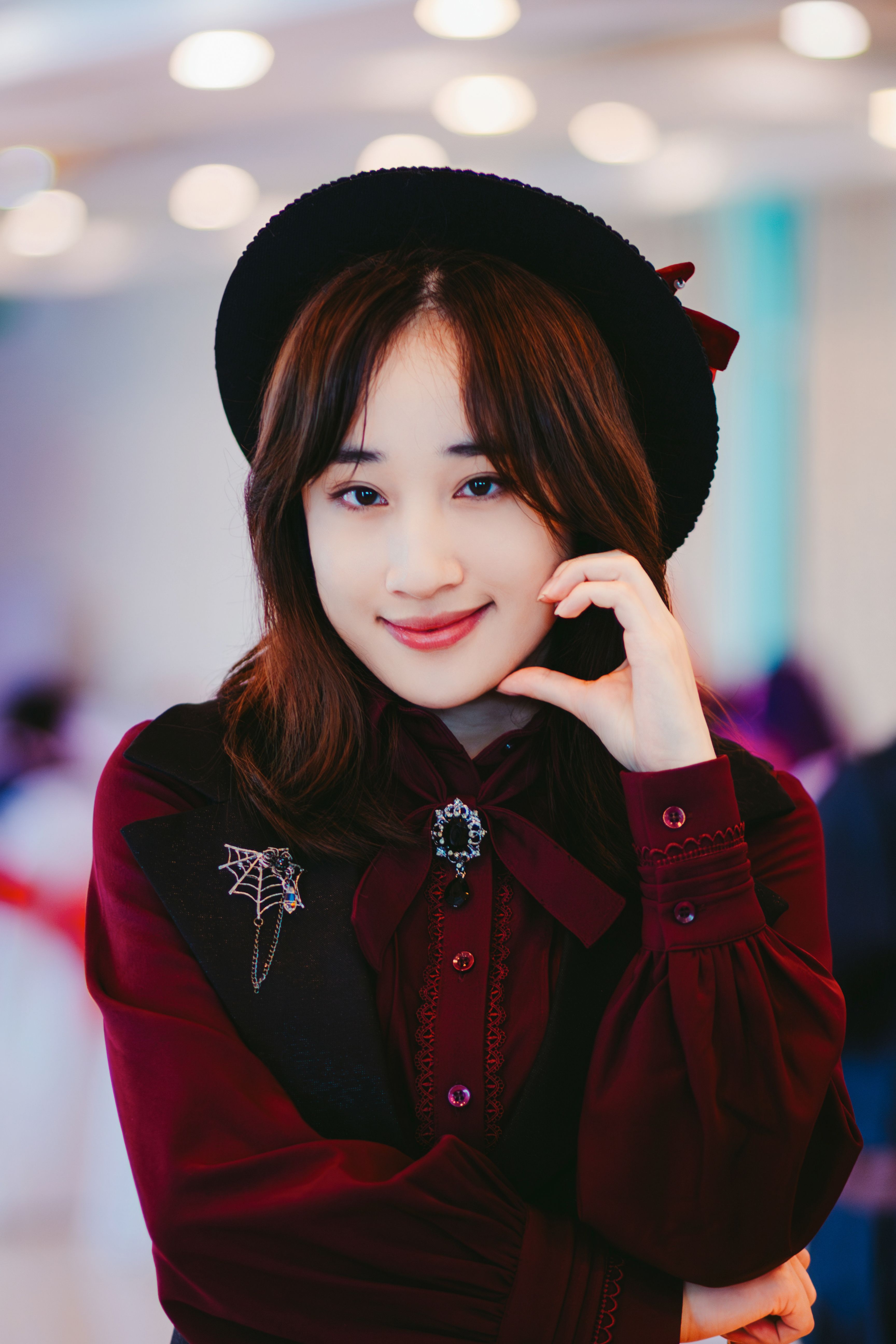 Smiling woman in cosplay attire with black hat and burgundy dress against a blurred indoor background.