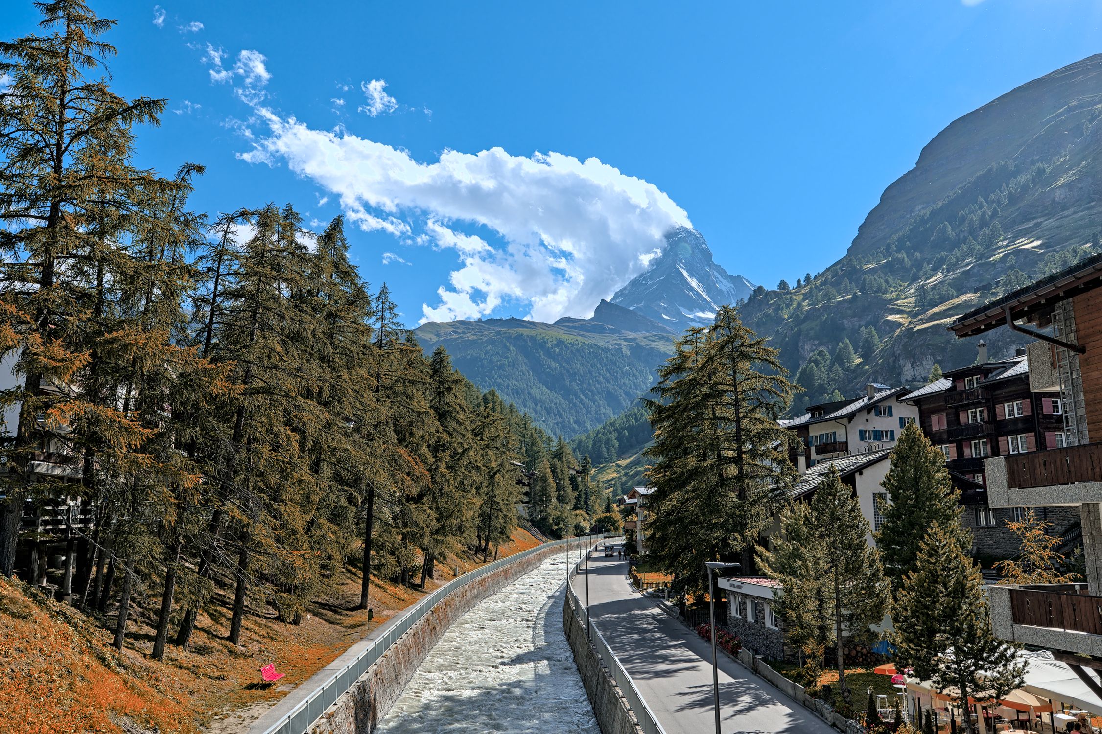 Discover the picturesque town of Zermatt with the iconic Matterhorn towering in the background, featuring lush trees and a flowing river.
