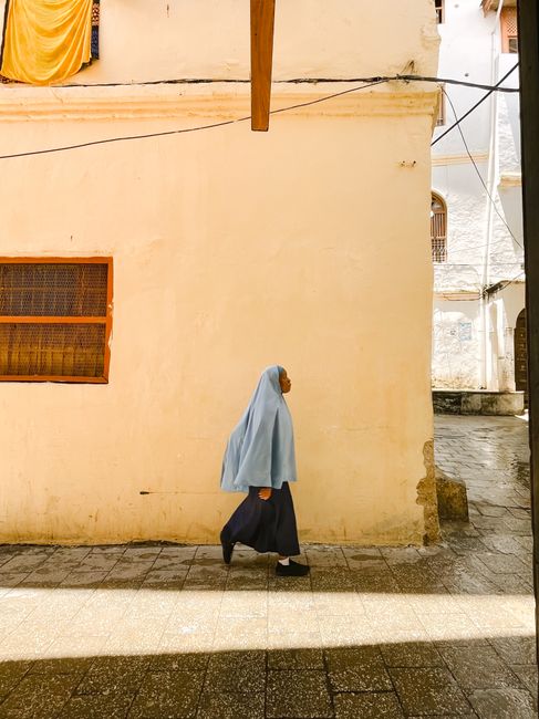 A woman in traditional clothing walks along a sunlit street in Zanzibar, Tanzania.