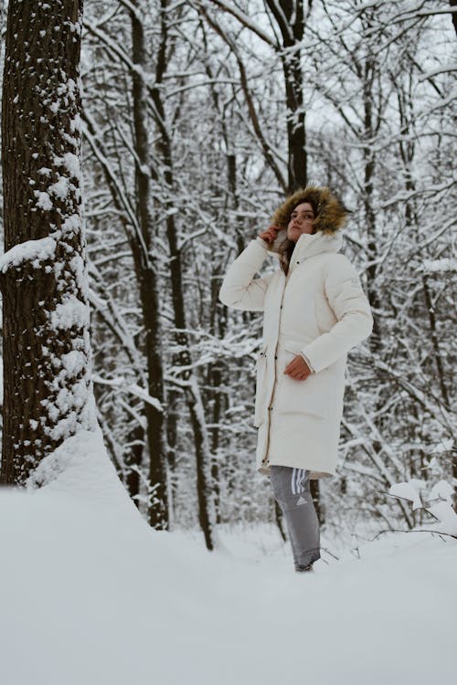 Model in a White Padded Winter Coat with a Fur-Trimmed Hood and Gray Adidas Tracksuit Pants Standing in a Snow Covered Forest