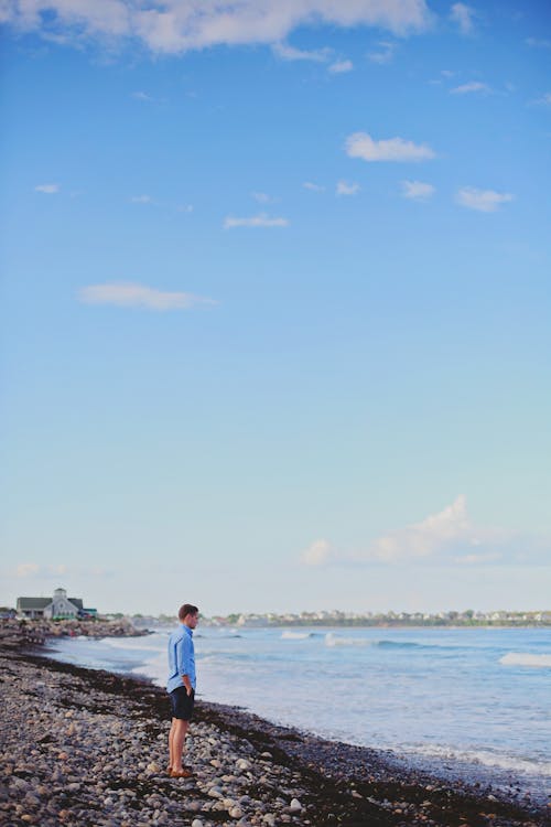 Gratis Pria Berdiri Di Tepi Pantai Di Depan Laut Di Bawah Langit Berawan Foto Stok