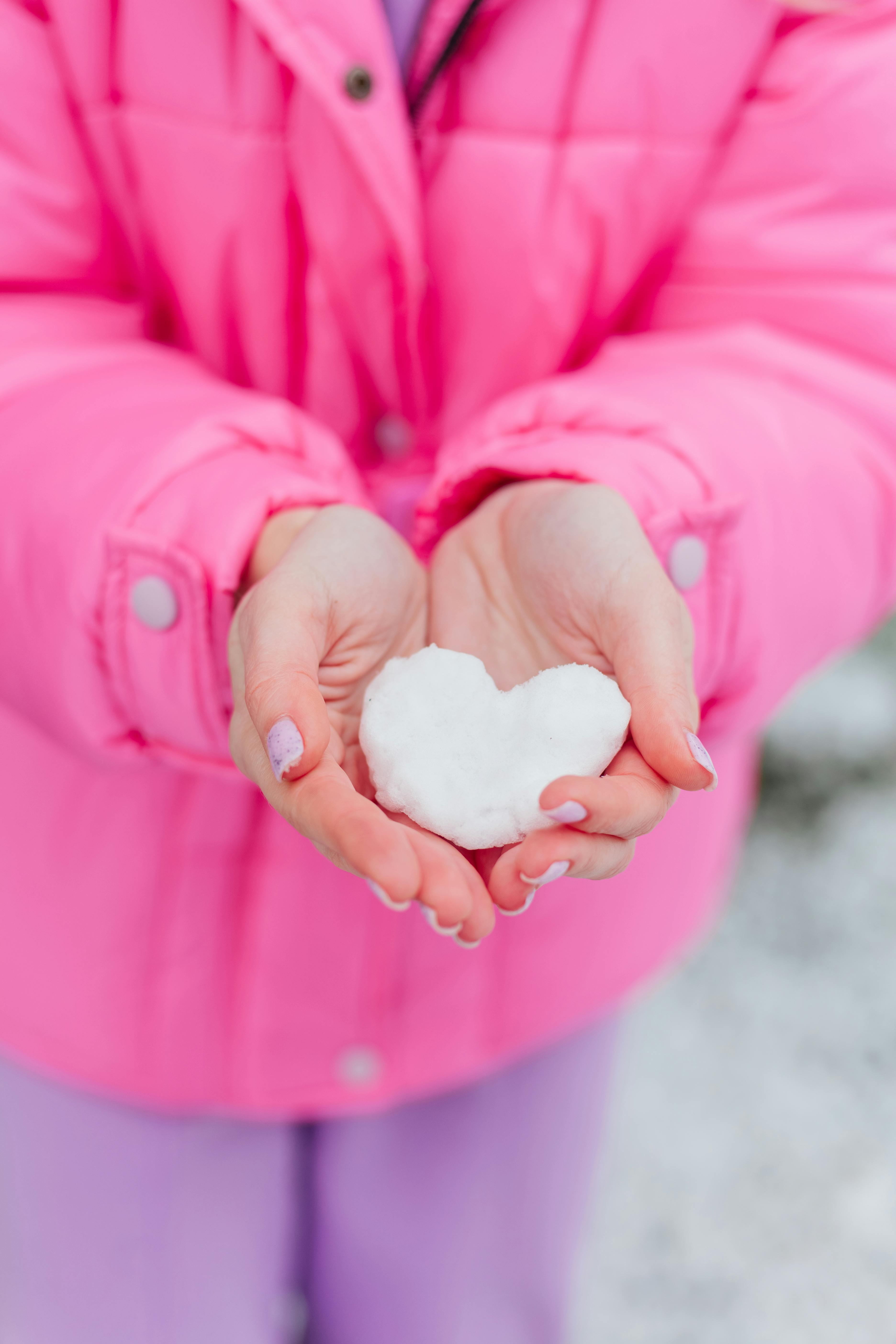 A young girl in a pink coat holds a heart-shaped snow piece, symbolizing love and winter fun.