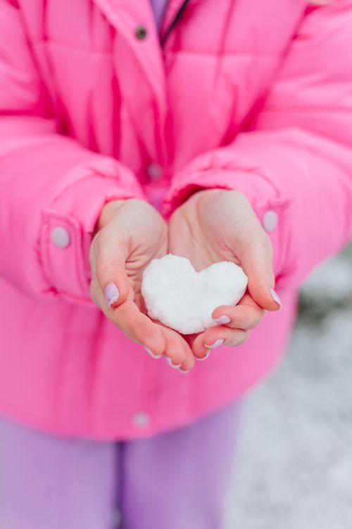 Free A young girl in a pink coat holds a heart-shaped snow piece, symbolizing love and winter fun. Stock Photo
