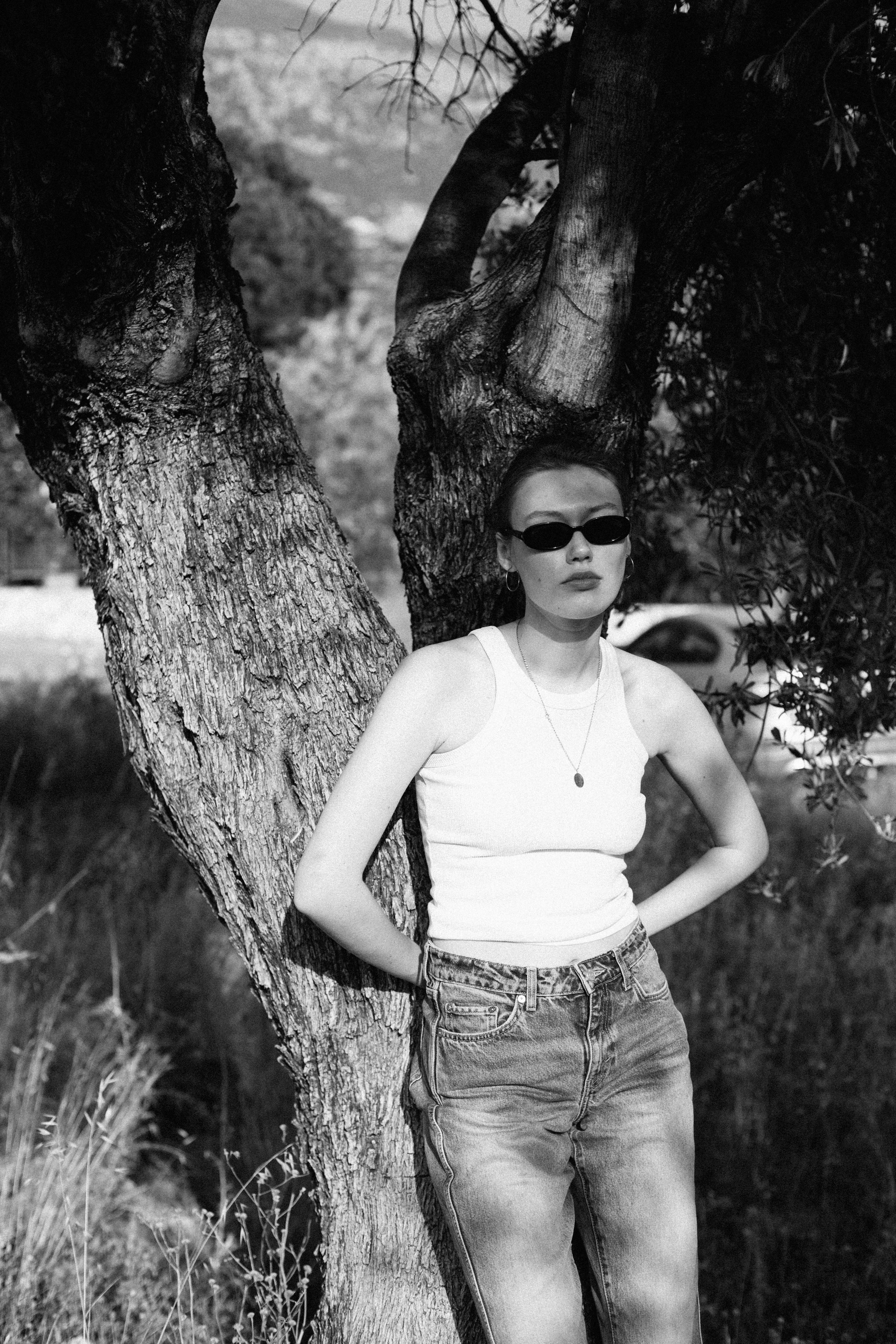 Chic woman in sunglasses leaning against a tree, shot in black and white.