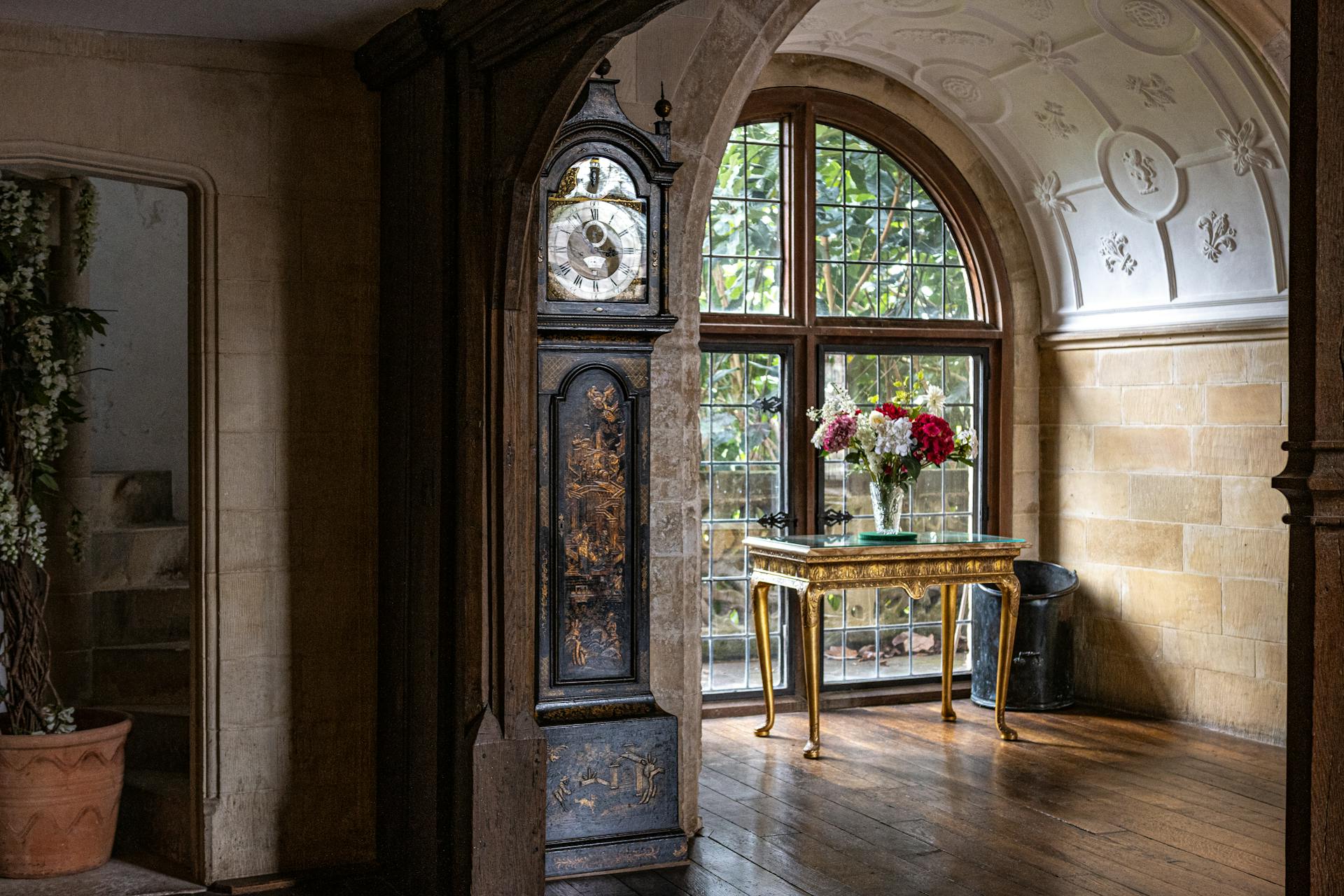 Elegant vintage room featuring an ornate grandfather clock and a floral-adorned table by a large window.