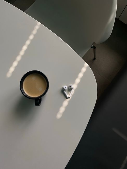 Overhead view of coffee cup and wireless earbuds on a table in soft morning light.