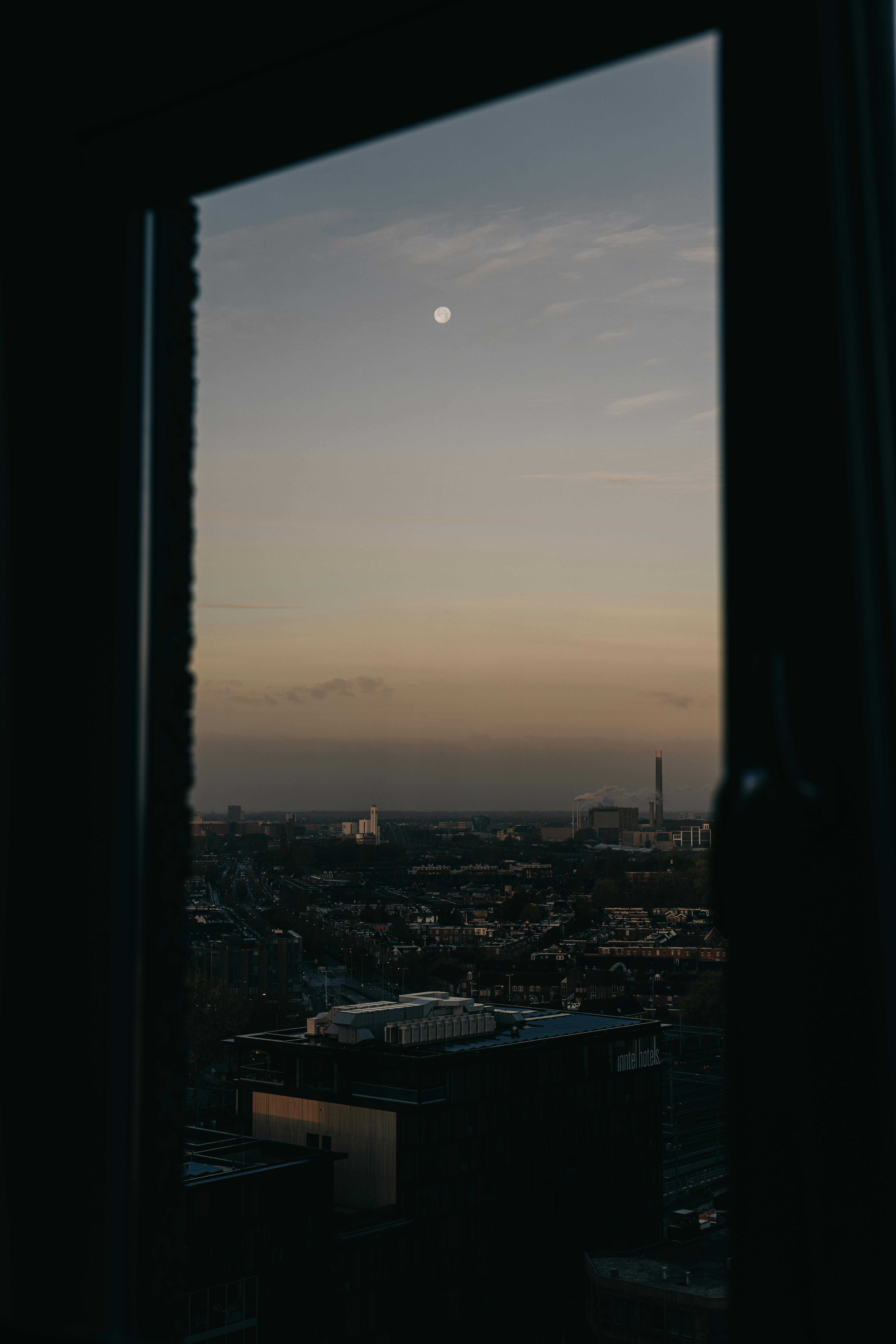 Serene cityscape view through a window with the moon rising at dusk, capturing urban tranquility.