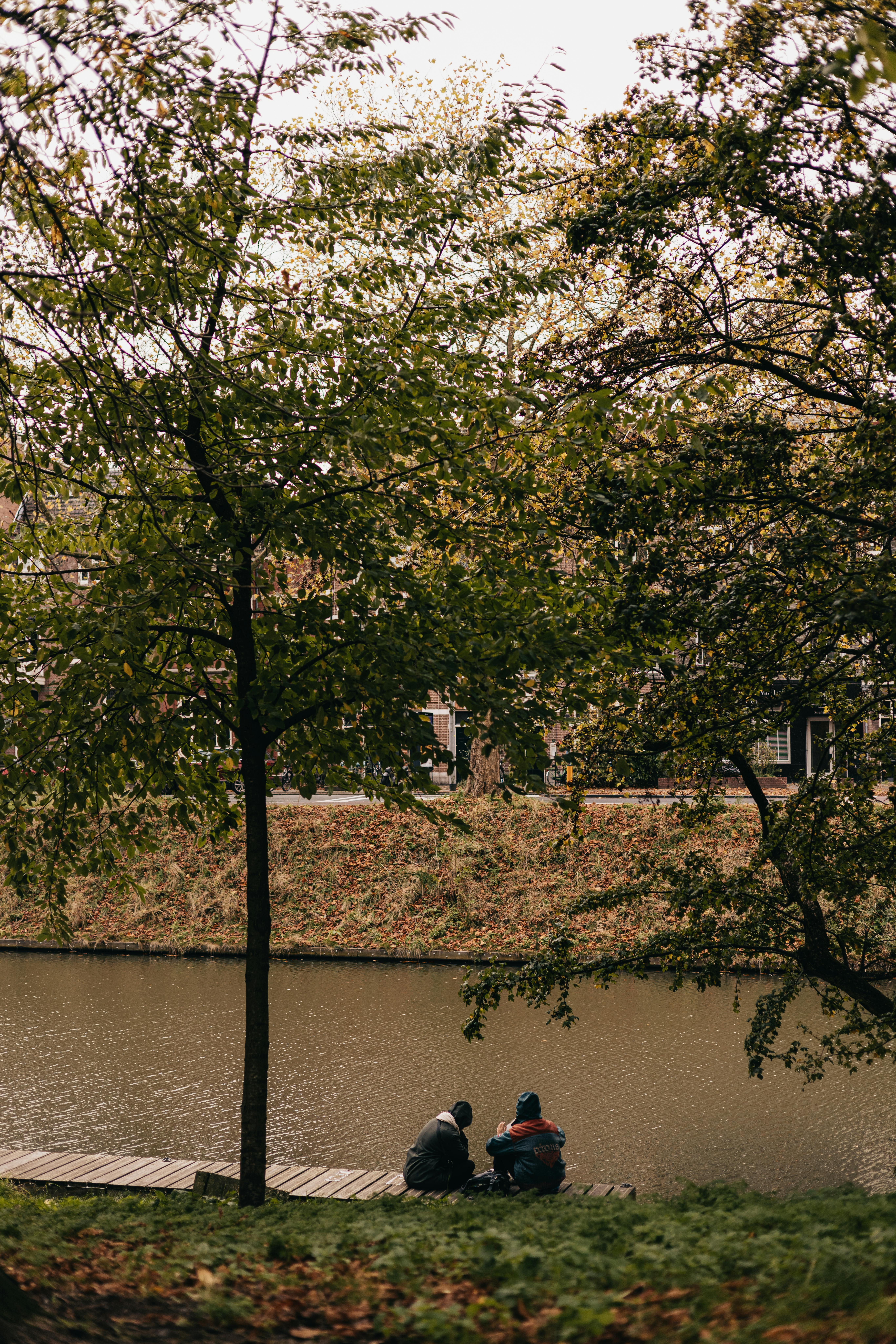 Two individuals sitting peacefully by a river under lush green trees during a cloudy day.
