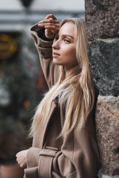 A stylish young woman poses in a brown coat against a stone wall, showcasing fashion and beauty.