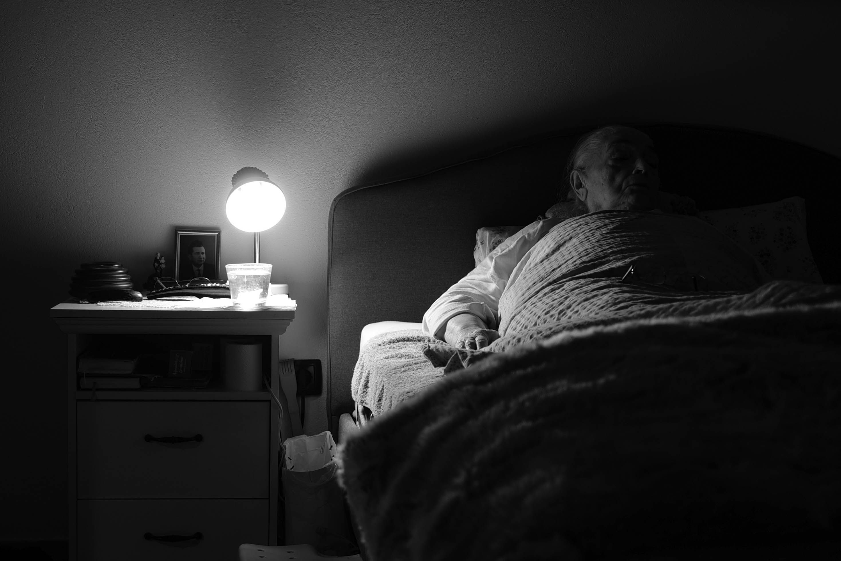 An elderly person resting in bed with a lit lamp beside in a cozy setting.