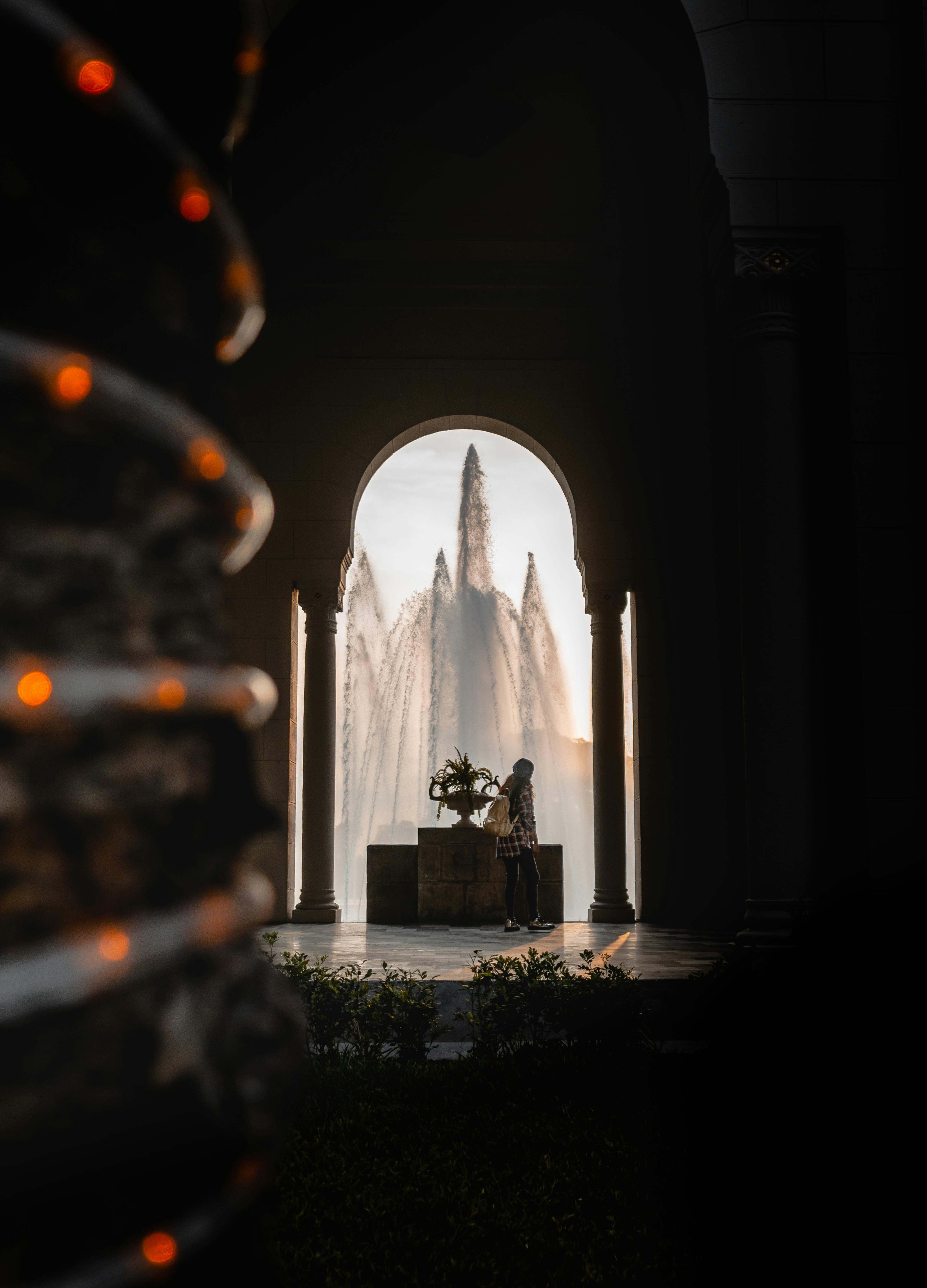A silhouette of a woman beside an ornate fountain framed by archways at sunset, creating a dramatic scene.