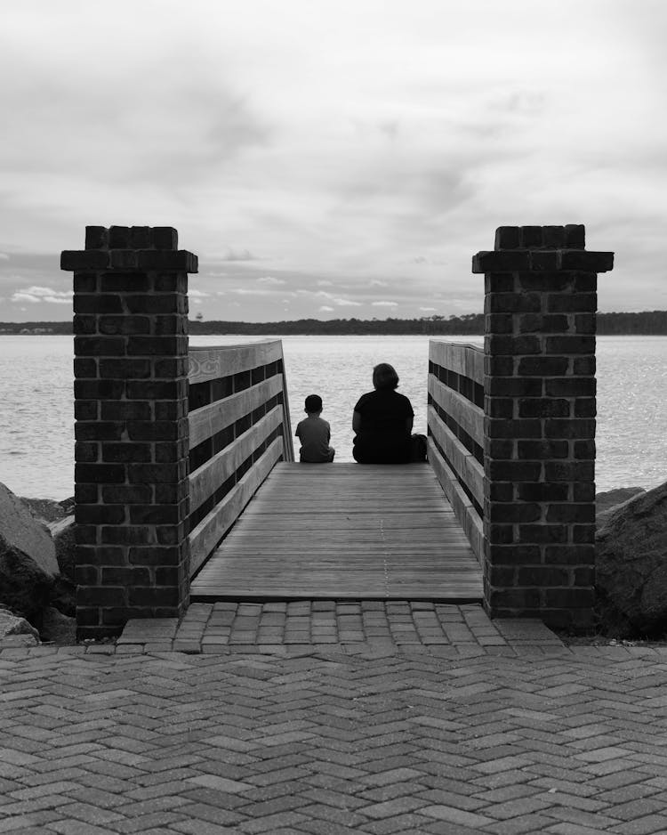 Back View Of An Adult And A Child Sitting On The Steps Of A Pier 