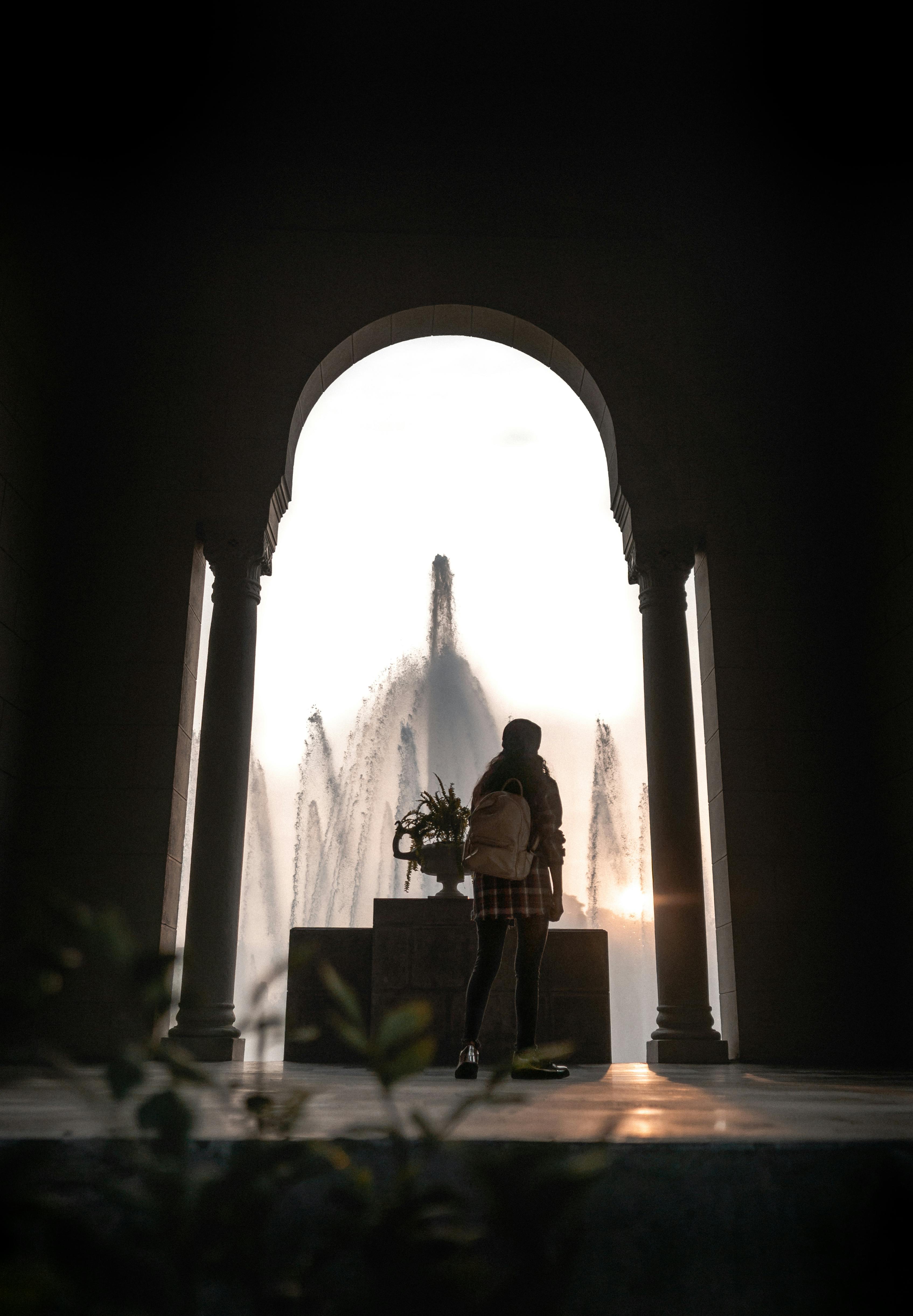 Silhouette of a young woman posing in front of a grand fountain during a sunset.