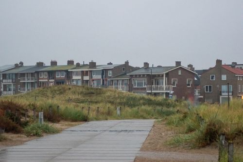 Free A serene view of a coastal residential street lined with grass-covered dunes on a cloudy day. Stock Photo