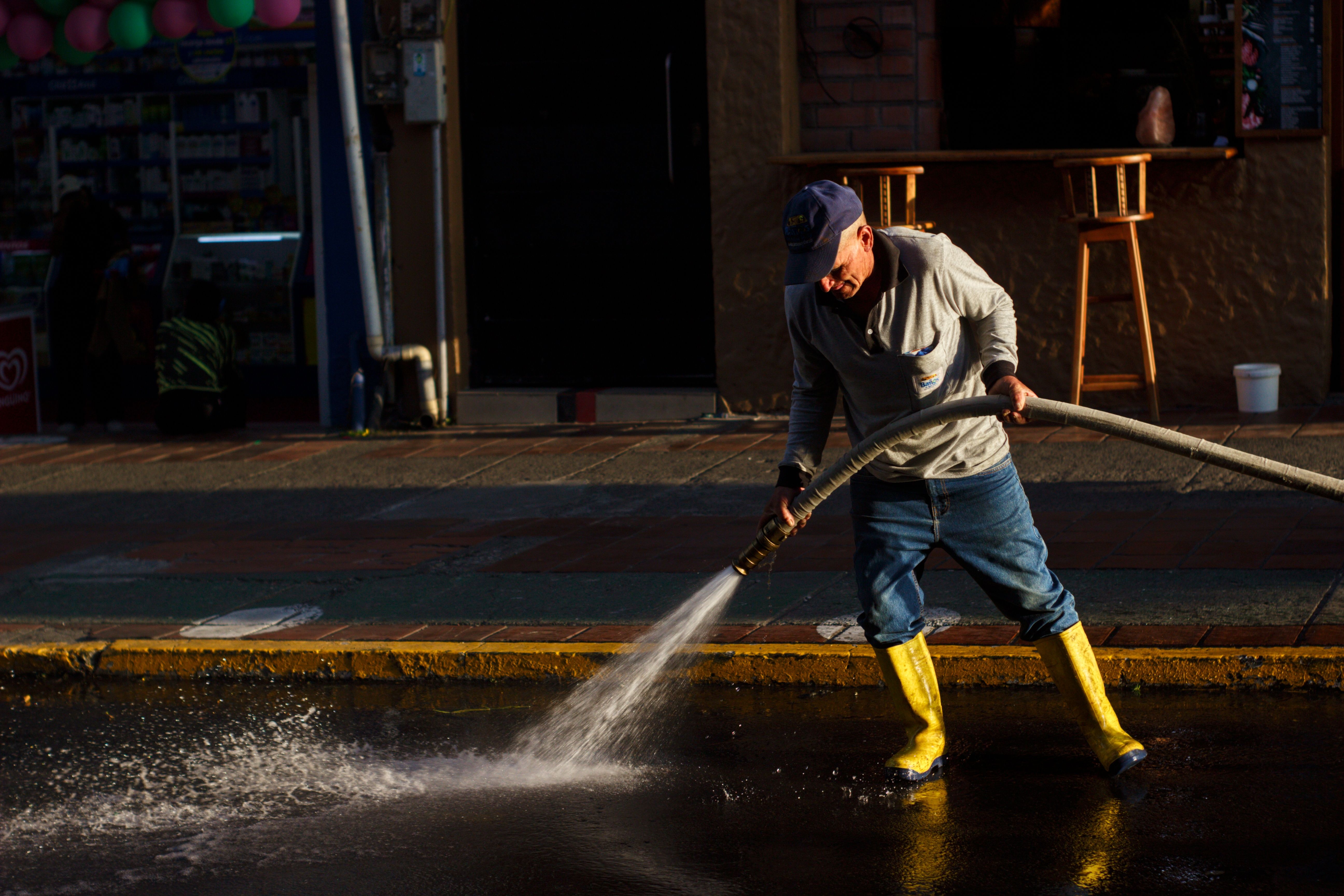 A man using a water hose to clean a city street, wearing yellow rain boots and a cap.
