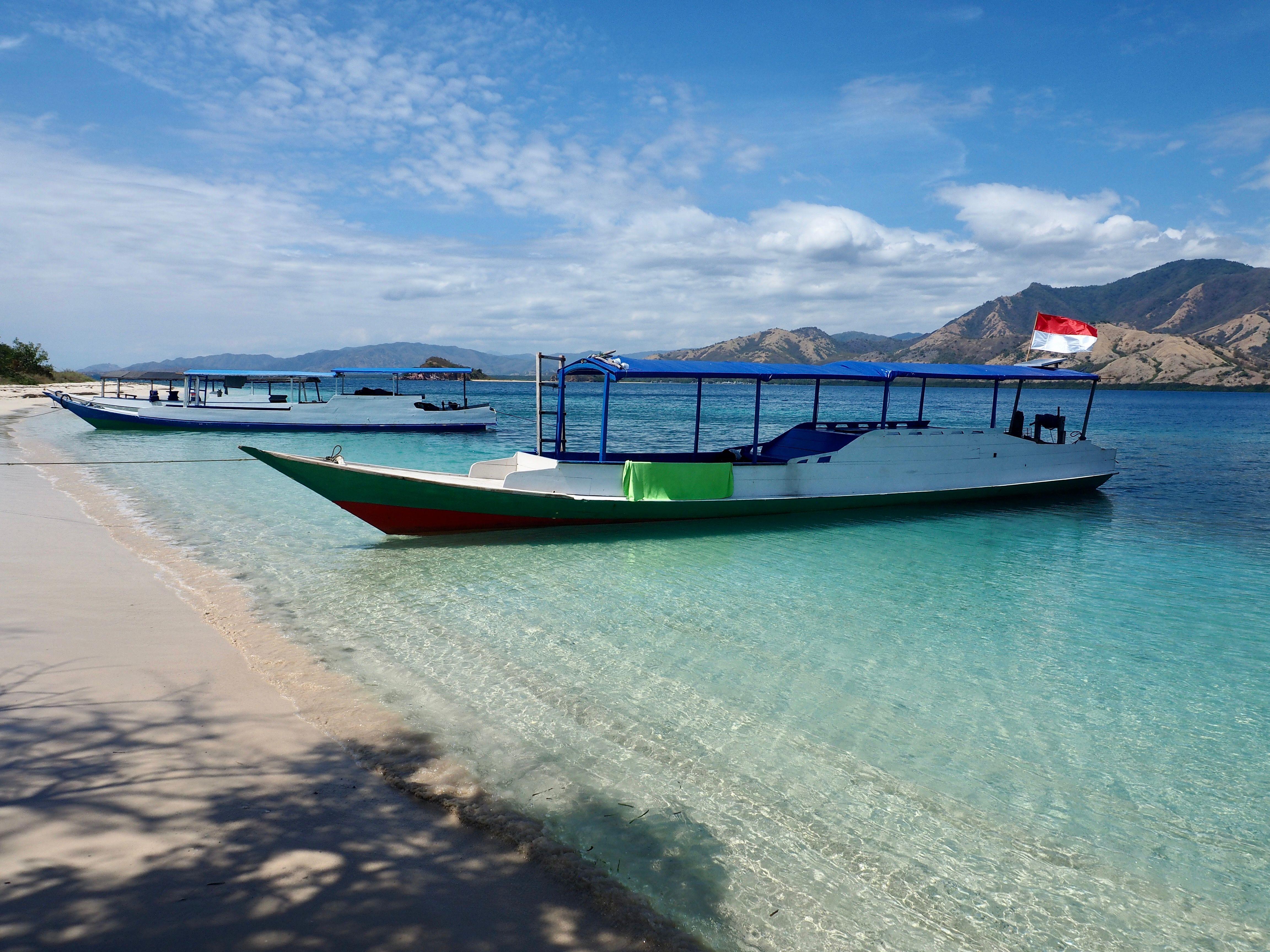 Traditional Indonesian boats moored at a serene tropical beach with clear blue waters and distant mountains.