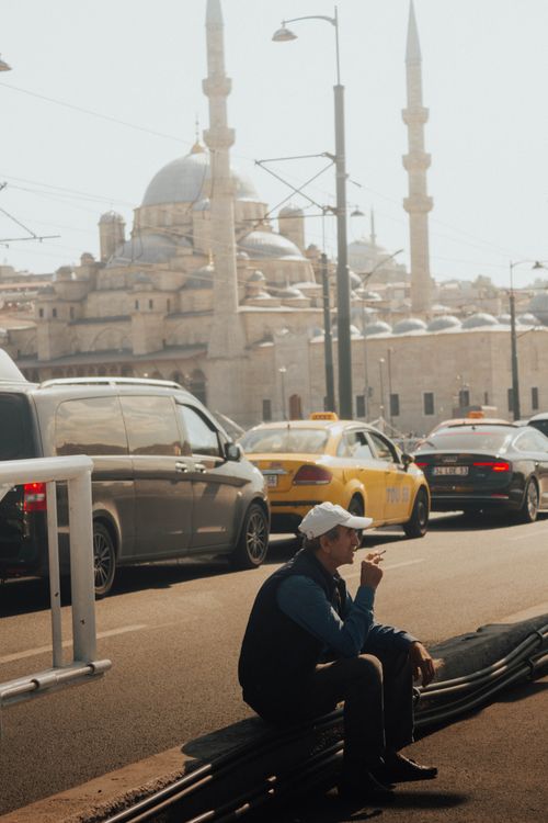 Free A man is sitting and smoking near a bustling street in Istanbul, with a mosque and traffic in the background. Stock Photo