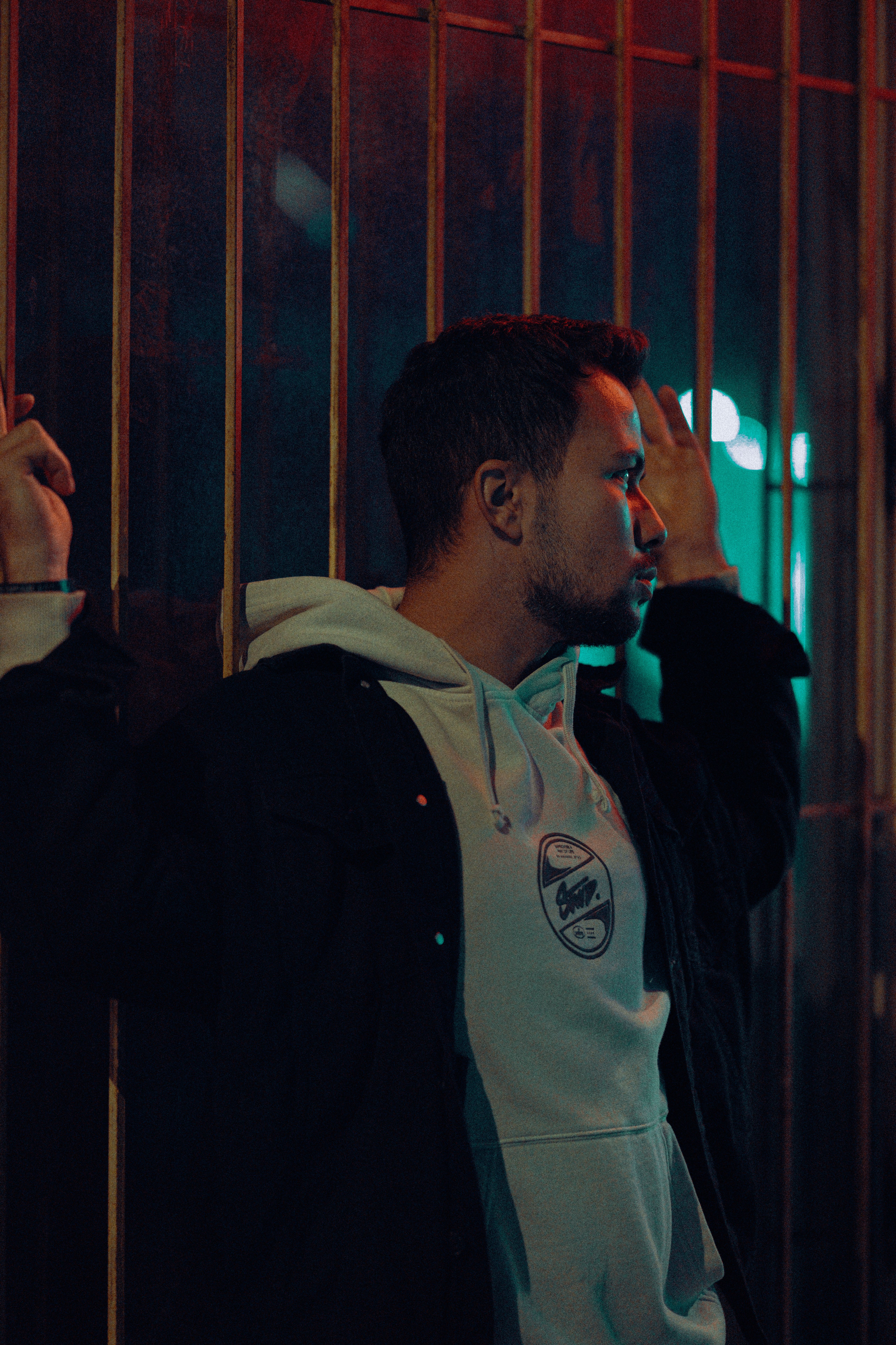 Portrait of a young man in a hoodie and jacket leaning against a fence at night in an urban setting.