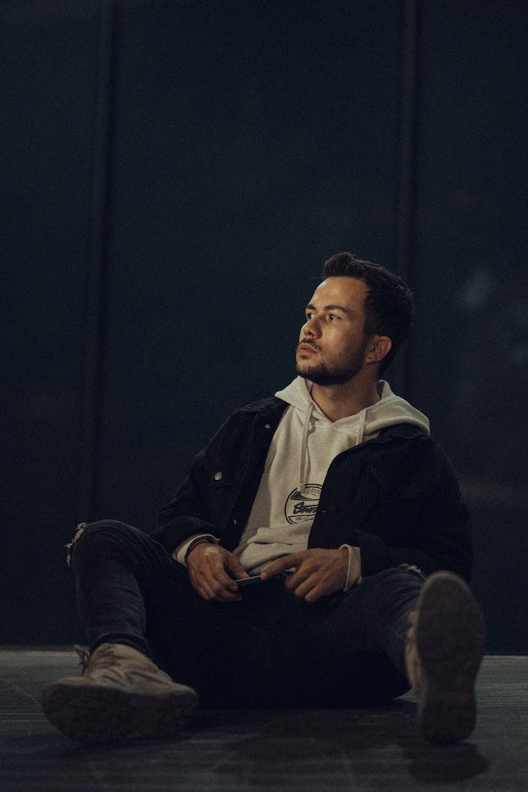 Man In Jacket And Hoodie Sitting On Ground At Night