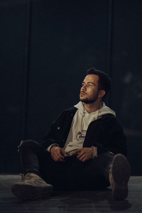 Free Portrait of a young man in a hoodie and jacket, sitting at night in an urban setting. Stock Photo