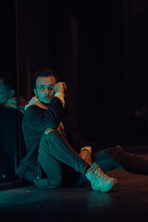 Free A man in a hoodie and jacket sits thoughtfully under low light indoors at night. Stock Photo